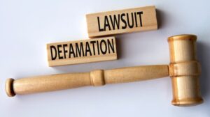 The phrase "DEFAMATION LAWSUIT" is displayed on wooden blocks next to a judge's gavel against a white background.